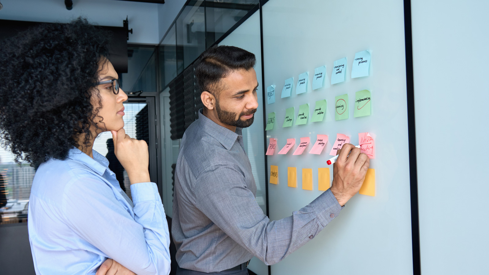 Young indian businessman manager writing strategy ideas on sticky notes on whiteboard and female African American colleague looking at his strategy scrum presentation. Business project planning.