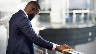 Stressed african american young man in suit entrepreneur standing by railing with head down, experiencing difficulties at work, back view shot, panorama with copy space, financial crisis and business