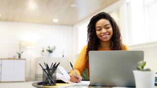 Smiling lady sitting at desk, using laptop and writing in notebook, taking notes, watching tutorial, lecture or webinar, studying online looking at screen, free copy space